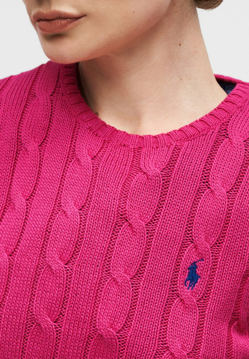 Femme portant un pull en maille torsadée rose vif avec col rond côtelé et petit logo brodé bleu marine sur la poitrine.