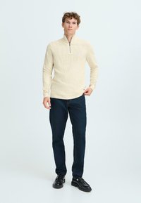 Licht beige gebreide pullover met een half-rits kraag, geribbelde textuur, slim-fit ontwerp, gecombineerd met donkerblauwe jeans en zwarte schoenen.