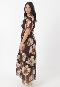Robe maxi en tissu transparent marron avec de grands motifs floraux en beige et crème. Présente des manches courtes et un design cintré à la taille.