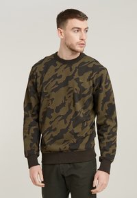 Sudadera de camuflaje en tonos verde oliva y negro, con cuello redondo y puños acanalados. La tela es una mezcla de algodón con una textura suave.