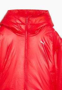 Rode pufferjack met een glanzende afwerking, volledige rits, hoge kraag en grote capuchon. Heeft een wit Puma-logo op de linkerborst.