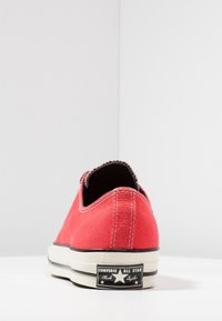 Baskets en toile rouges au design bas, semelle en caoutchouc blanc, bord noir et une étiquette avec "Converse All Star". Surface texturée.