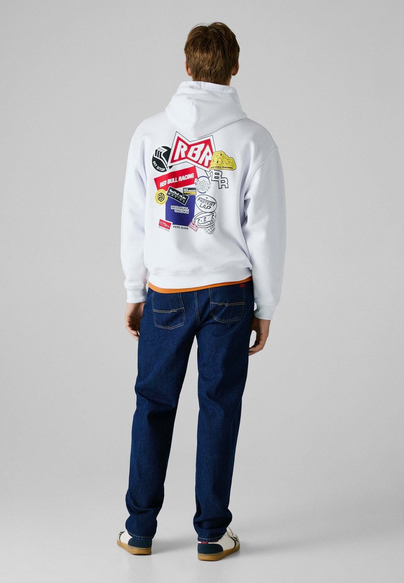 Felpa bianca con cappuccio caratterizzata da vari patch grafici, inclusi loghi. Abbinata a jeans blu scuro e scarpe casual, modello visto da dietro.