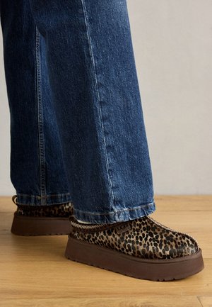 Des chaussures à enfiler à motif léopard avec une semelle épaisse marron, associées à un jean large bleu, reposant sur un sol en bois.