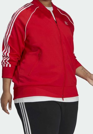 Osoba w czerwonej kurtce sportowej Adidas z białymi paskami i czarnych spodniach z białymi paskami, stojąca na jednolitym tle.
