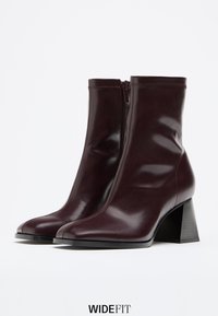 Stivali alla caviglia in pelle bordeaux con punta quadrata, cerniera frontale e tacco a blocco, caratterizzati da una texture liscia e un design minimalista.