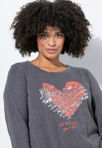 Maglietta grigia a maniche lunghe con un design a cuore in paillettes corallo e argento. Il testo recita "DIFFONDI il tuo amore" sotto il grafico. Materiale morbido, vestibilità casual.