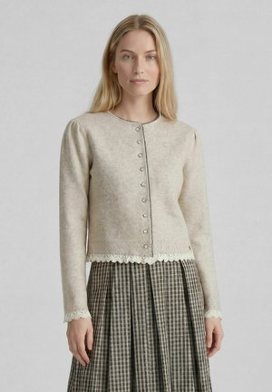Frau mit glatten blonden Haaren, die eine beige, geknöpfte Strickjacke mit gezackten Rändern und einen plissierten karierten Rock trägt.