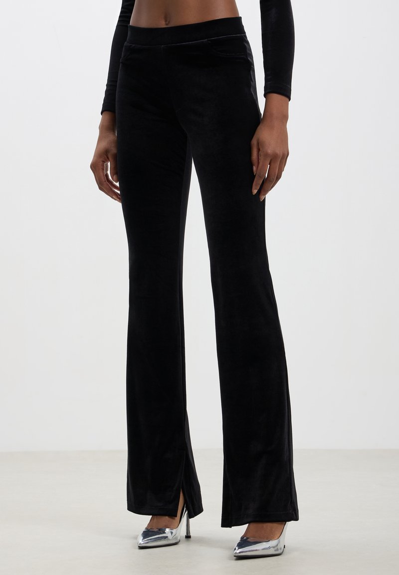 Femme portant un pantalon évasé en velours noir avec des fentes sur le devant, associé à des talons hauts pointus argentés sur fond uni.