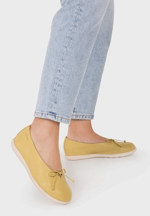 Osoba nosząca jasnoniebieskie dżinsy i żółte skórzane baleriny z małymi kokardkami i białymi podeszwami.
