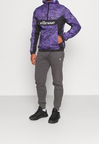 Lila kamouflage vindtäktare med svarta detaljer och logotyp, parat med gråa träningsoveraller. Svarta sportskor fulländar outfiten.