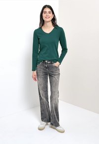 Grünes langärmeliges V-Ausschnitt-Oberteil mit glatter Textur, kombiniert mit grauen geraden Jeans und weißen Sneakers mit beige Akzenten.