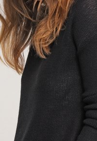Černý pletený svetr s texturovaným povrchem a širokým výstřihem. Rukávy jsou dlouhé a přiléhavé, což podtrhuje uvolněný styl.