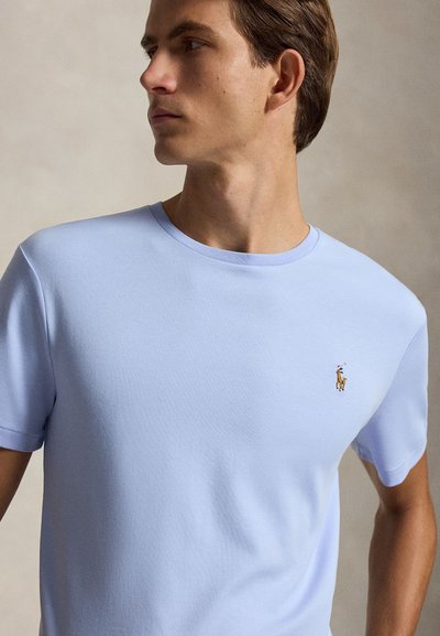 Jeune homme portant un T-shirt bleu clair à col rond avec un petit logo brodé d’un joueur de polo sur la poitrine gauche, regardant sur le côté.
