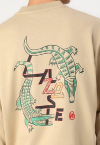 Smėlinis džemperis su daugiakompleksiu dviejų krokodilų grafiku, turinčiu geometrinius raštus ir ryškius tekstinius elementus ant nugaros.
