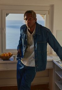 Traper jakna s prednjim gumbima i džepom na prsima, nošena preko svijetle košulje. Muškarac stoji kraj prozora, s narančama u zdjeli u blizini.