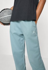 Pantaloni della tuta azzurri chiari con tasche e vita elastica, con il logo Jumpman bianco. T-shirt nera e pallone da basket in mano.
