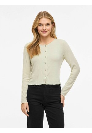 Ung kvinde med langt blondt hår, iført en lysebeige ribstrikket cardigan med knapper og sorte bukser med høj talje, stående med hånden på hoften.