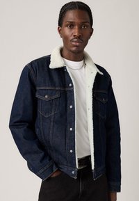 Chaqueta de denim con un exterior azul oscuro y forro de felpa blanca. Presenta botones a presión, dos bolsillos en el pecho y costuras en contraste.
