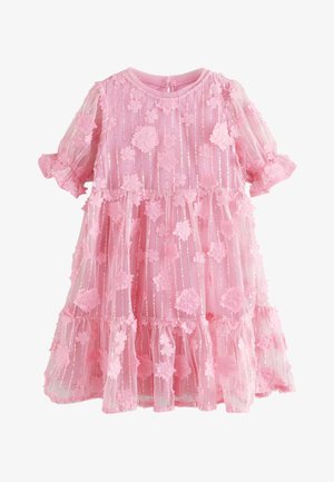 Ragazza giovane con capelli scuri intrecciati e fermaglio floreale rosa, che indossa un vestito rosa con applicazioni floreali, mentre tiene delicati fiori rosa vicino al volto.