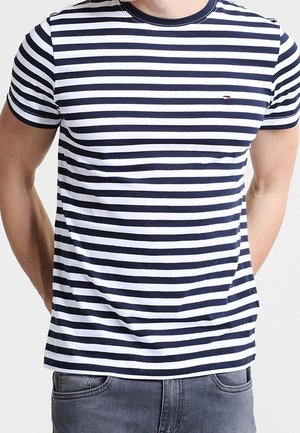 Uomo che indossa una maglietta a maniche corte a righe orizzontali blu navy e bianca e jeans grigi, in piedi con le mani dietro la schiena.