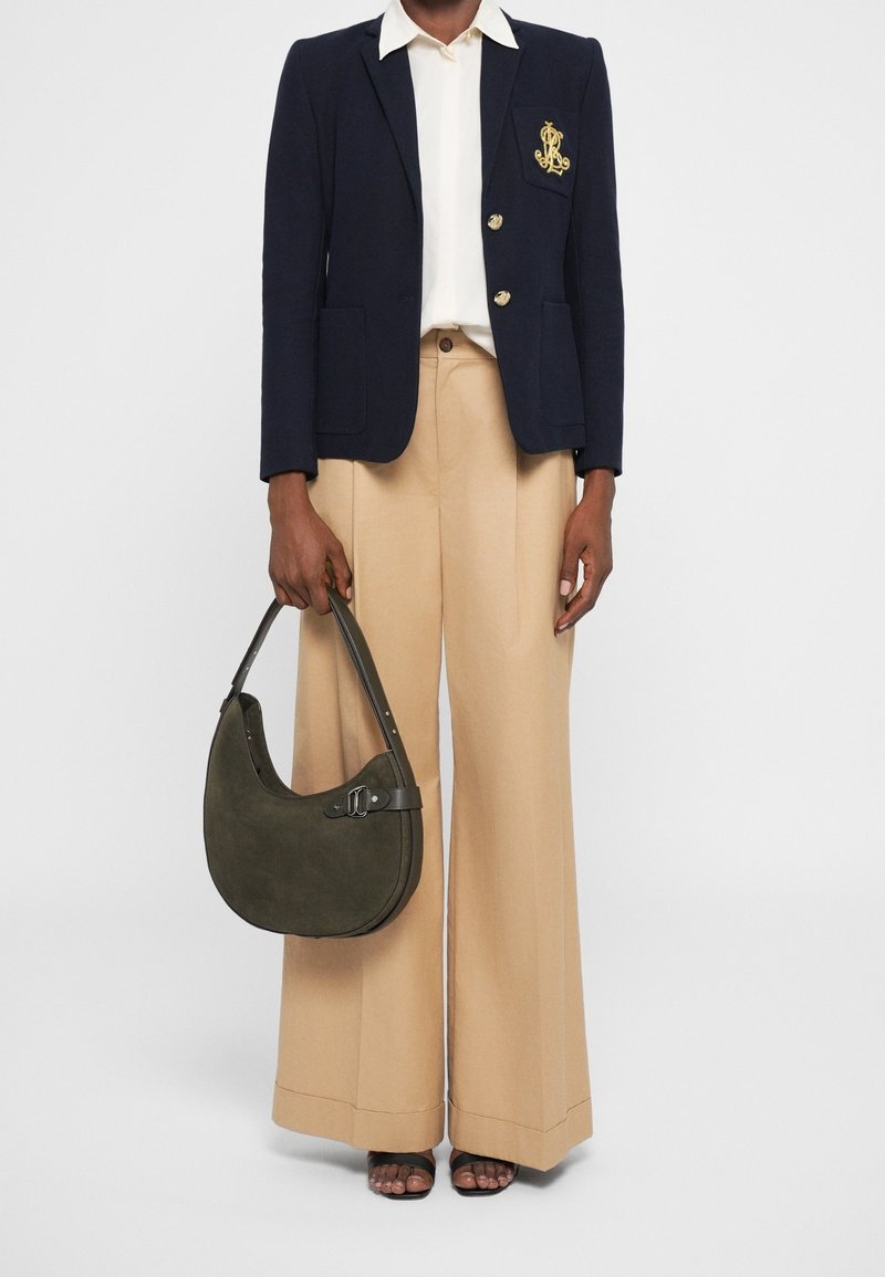 Personne portant un blazer bleu marine, une chemise blanche, un pantalon large beige, des talons ouverts noirs, tenant un sac vert olive en forme de croissant.