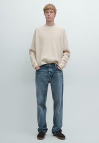 Un giovane con pelle chiara e capelli rossi, che indossa un maglione a coste crema, jeans blu larghi e scarpe marroni scure, in piedi con le mani in tasca.