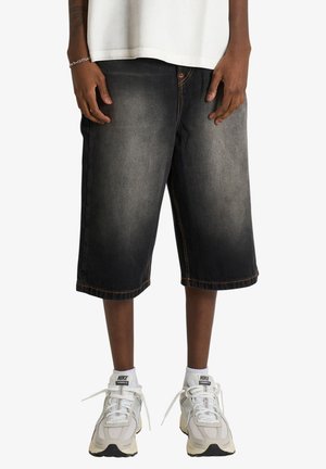 Persona che indossa pantaloncini di jeans neri sbiaditi alla lunghezza del ginocchio, scarpe da ginnastica Nike bianche con calzini bianchi e una maglietta bianca, in piedi con le mani rilassate.