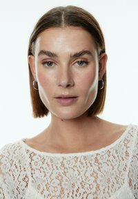 Model mit kurzen, glatten braunen Haaren, trägt ein weißes Spitzenoberteil mit Blumenmustern und einem Bootsausschnitt. Silbern Ohrringe in Form von Ringen sind sichtbar.