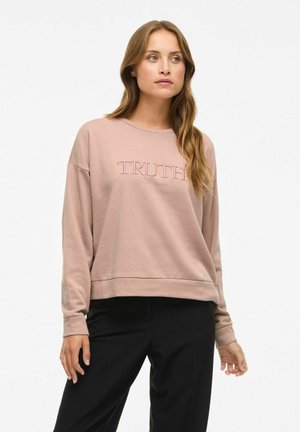 Sweatshirt rose clair avec le mot "VÉRITÉ" brodé dans une teinte contrastante. Présente une coupe décontractée et des manches longues. Associé à un pantalon noir.