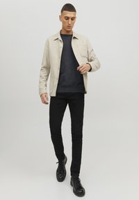 Chaqueta beige de botón sobre un suéter oscuro, combinada con jeans ajustados negros y botas negras con cordones, con un bolsillo en la chaqueta.