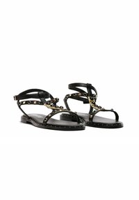 Chanclas de cuero negro con tachuelas y acentos dorados. Suela plana, diseño de punta abierta, banda única alrededor del tobillo, detallado recortado intrincado.