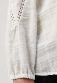Hvit teksturert bluse med semi-transparent stoff, med subtile horisontale striper og pyntet med blonderdetaljer på ermene.