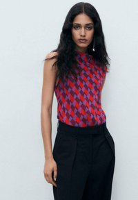 Short-sleeve top featuring a geometric pattern in red and purple tones, paired with high-waisted black trousers. Smooth fabric texture.