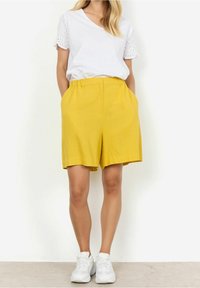 Żółte lniane szorty z elastycznym paskiem i bocznymi kieszeniami, zestawione z białą koszulką z koronkowymi akcentami na rękawach.