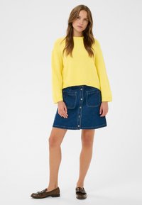 Pull en maille jaune à large encolure, associé à une jupe trapèze en denim foncé avec poches avant et fermeture par boutons. Mocassins marron.