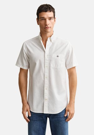 Man wearing a white short-sleeve button-up shirt with chest pocket logo and blue jeans, standing against a plain white background.