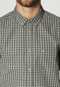 Koszula na guziki w zielono-szary wzór w kratę. Posiada kołnierzyk, brązowe guziki oraz małe wyszywane logo na lewej kieszeni na piersi.