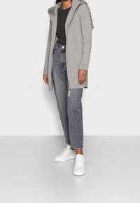 Felpa grigia con cappuccio e chiusura con zip, abbinata a jeans grigi a vita alta e sneaker bianche. Tessuto morbido, vestibilità leggermente ampia.