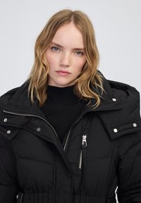 Giacca nera imbottita con colletto alto, zip argento, bottoni a pressione e vita elasticizzata. Tessuto liscio con texture trapuntata.