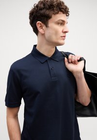 Camisa polo azul marinho de mangas curtas, com uma pala de três botões e logótipo no peito. O modelo transporta uma bolsa tote preta sobre um dos ombros.
