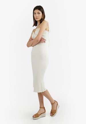 Donna in piedi con le braccia incrociate, indossa un abito midi crema senza maniche e sandali marroni con zeppa su sfondo bianco.