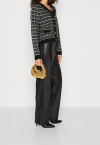 Cardigan in tweed bianco e nero con grandi bottoni, abbinato a pantaloni larghi in pelle nera. Tiene in mano una piccola borsa a rete dorata.