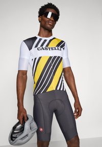 Kortermet sykkeltrøye i hvitt, svart og gult med stripet mønster; kombinert med grå shorts. Modellen holder en grå hjelm.