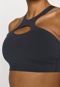 Μαύρο αθλητικό σουτιέν με σχέδιο με ανοίγματα και φαρδιές τιράντες. Κατασκευασμένο από απαλό ύφασμα με ραμμένες λεπτομέρειες και μικρό λογότυπο στο μπροστινό μέρος.