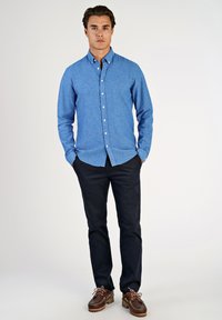 Camicia azzurro chiaro con bottoni e collo, abbinata a pantaloni scuri e scarpe marroni. La camicia ha maniche lunghe e orlo dritto.