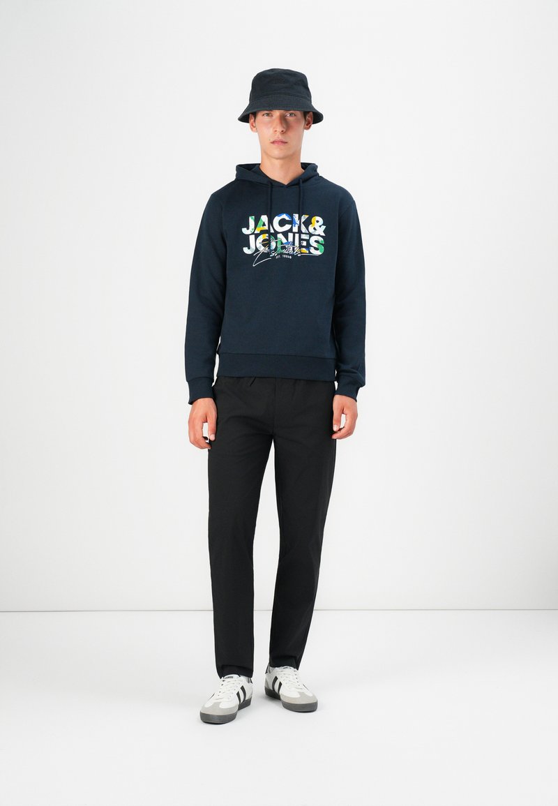 Marineblaues Hoodie mit "JACK & JONES"-Logo und bunten Akzenten, kombiniert mit schwarzen Hosen und weißen Sneakers mit schwarzen Streifen. Schwarzer Bucket Hat.