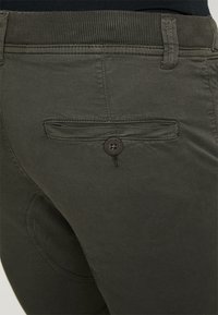 Pantalones de algodón verde oscuro con una cinturilla acanalada, un bolsillo trasero con botón y una textura suave. Detalles de costura sutiles visibles.