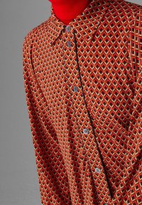 Rood en oranje gestreept shirt met geometrisch ontwerp, voorzien van een kraag, knopen en contrasterende zwarte accenten. Glad stoftextuur.