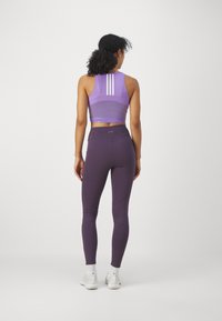 Top corto deportivo de color púrpura con tres franjas blancas en la parte posterior, combinado con leggins de color púrpura oscuro de cintura alta y zapatillas deportivas blancas.